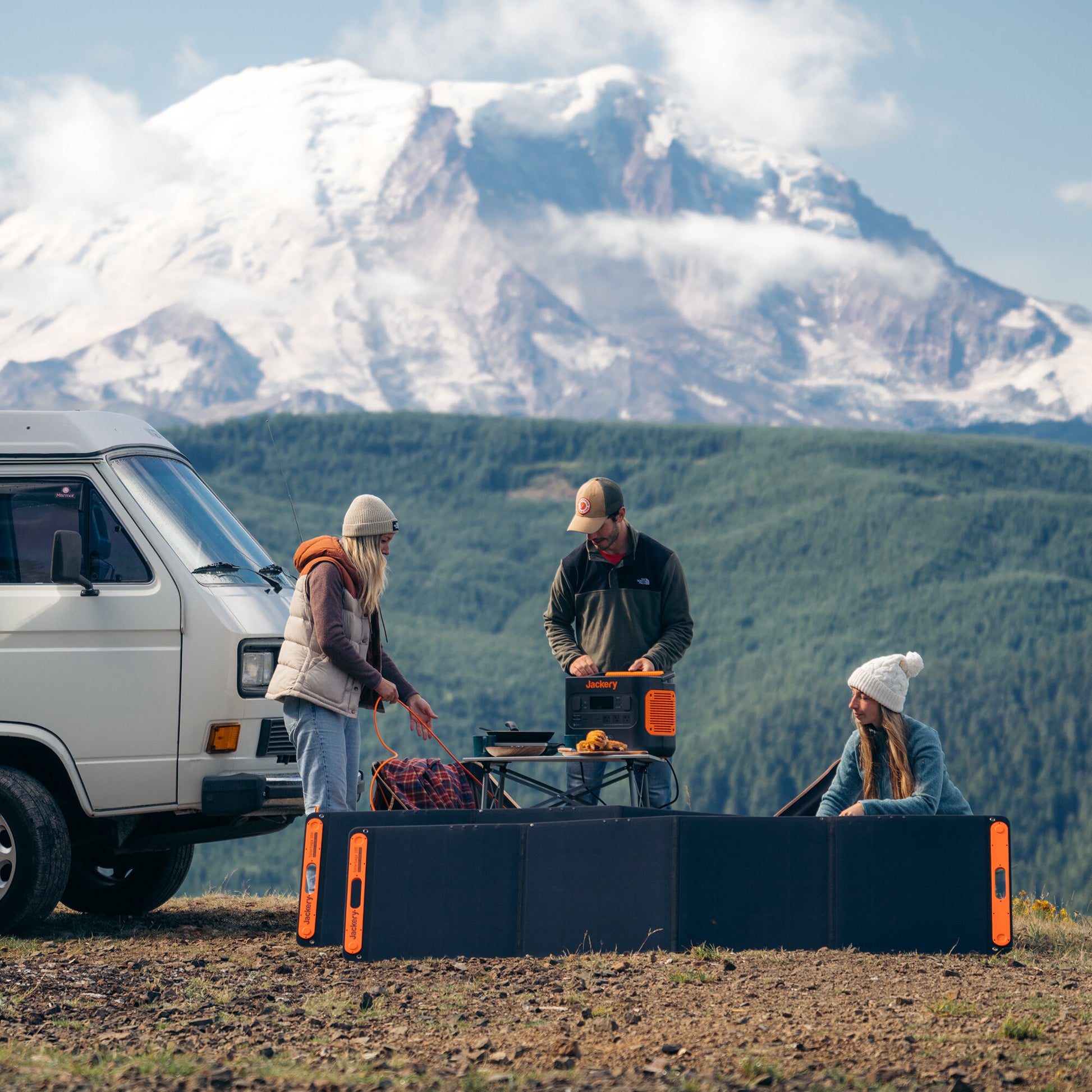 Two SolarSaga 200W panels unfolded on the ground beside a camper van with people cooking at a table