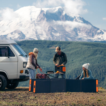 Two SolarSaga 200W panels unfolded on the ground beside a camper van with people cooking at a table