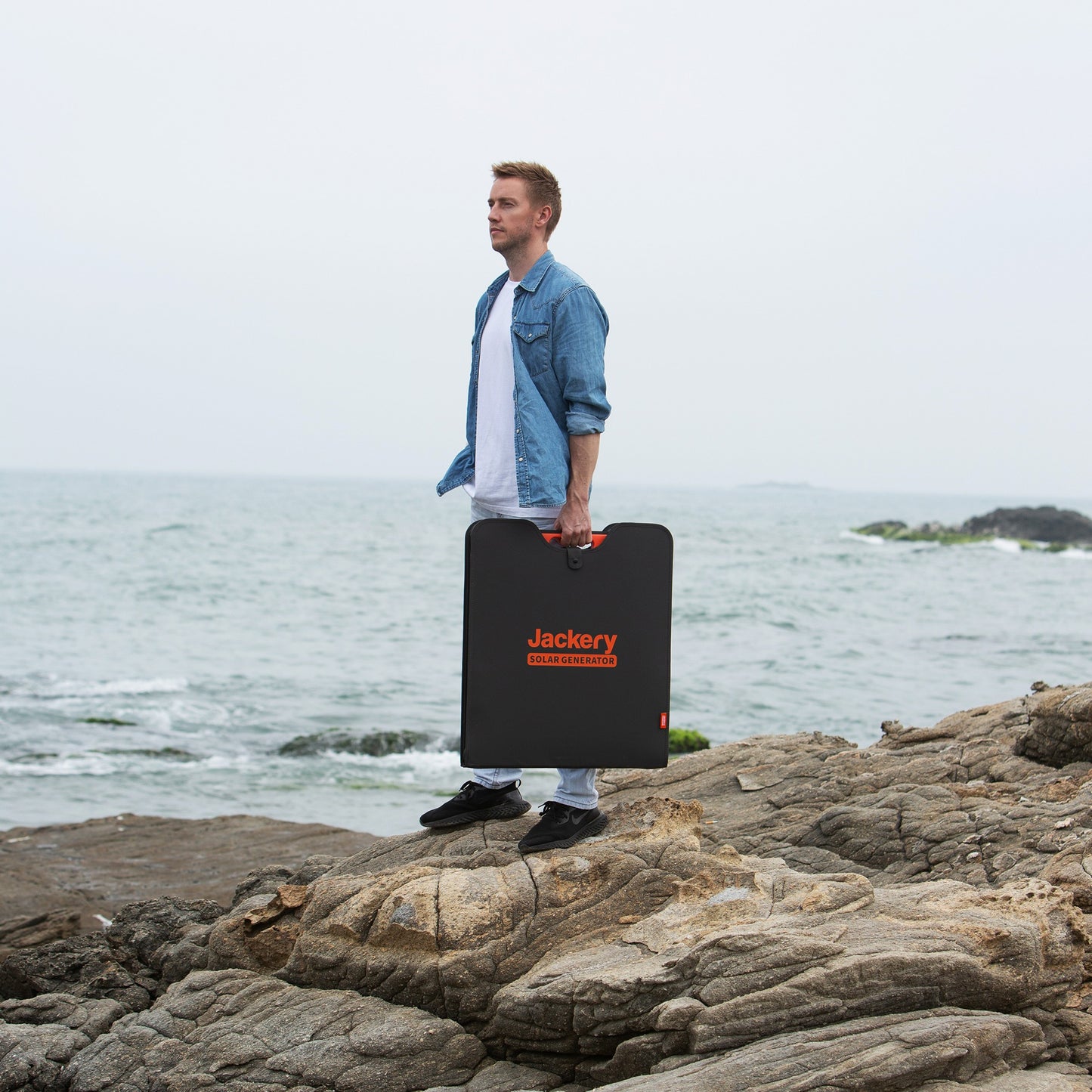Man carrying a folded SolarSaga 200W solar panel by its handle across coastal rocks