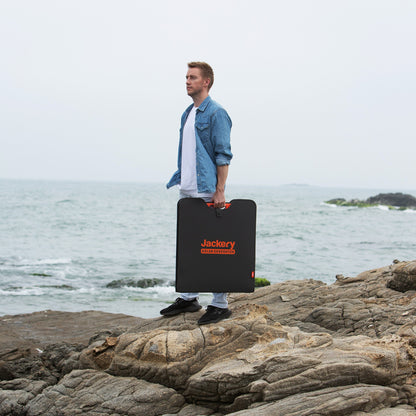 Man carrying a folded SolarSaga 200W solar panel by its handle across coastal rocks